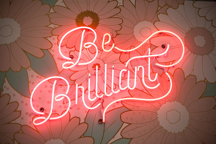 A word-based neon-light spelling out the words: "Be Brillaint" over flowery wall paper