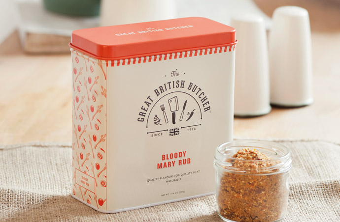 Photograph of the Bloody Mary Rub tin from the front with the lid on. The tin is rectangular shaped and has the Great British Butcher logo on the front and a pattern on the side made from repeating illustrations of the product ingredients. The tin is cream and the pattern and lid is dark green. The tin is situated in a traditional looking setting, with a ramekin containing the rub at the front and a traditional ceramic salt and pepper pot in the background.