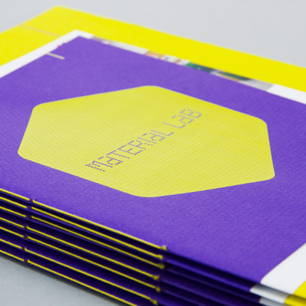 A stack of 7 Material Lab 20 page booklets. The cover stock is Colorplan Matrix emboss, in purple. On the cover is a large yellow screen printed hexagon with the Material Lab logo inverted in the centre. The sides of the booklet are visible and are saddle stitched with yellow thread.